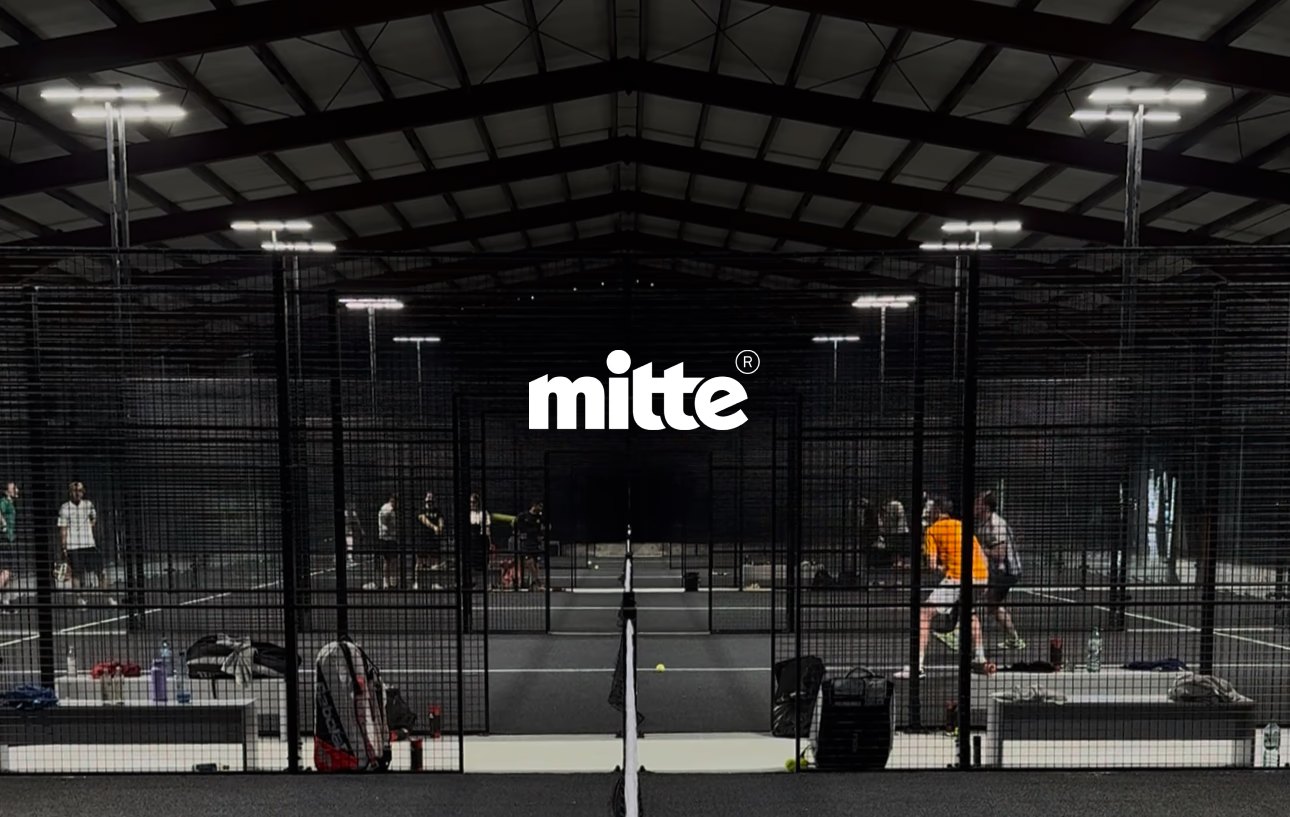 Blick in die Padelhalle von mitte: mehrere eingezäunte Courts unter einem Hallendach, im Hintergrund spielende Doppel